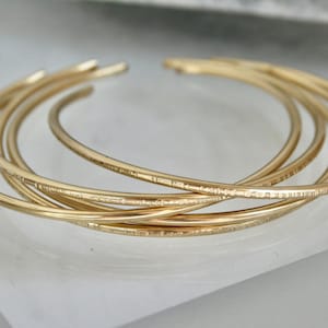 May include: A collection of gold-colored bangles. The bracelets are thin, round, and have a textured pattern. They are stacked and intertwined, creating a layered effect. The jewelry is displayed on a white surface.