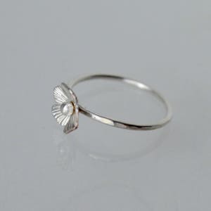 May include: A silver ring featuring a small, textured flower-shaped charm with a central bead. The band is thin and has a hammered finish, creating a reflective surface. The flower petals are detailed with fine, radiating lines.