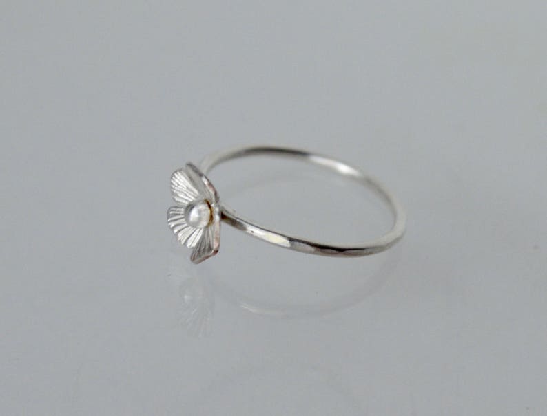 May include: A silver ring featuring a small, textured flower-shaped charm with a central bead. The band is thin and has a hammered finish, creating a reflective surface. The flower petals are detailed with fine, radiating lines.