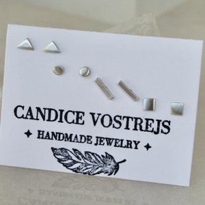 May include: A set of six silver stud earrings in various geometric shapes, including triangles, circles, and squares. The earrings are displayed on a white card with the text "CANDICE VOSTREJS HANDMADE JEWELRY".