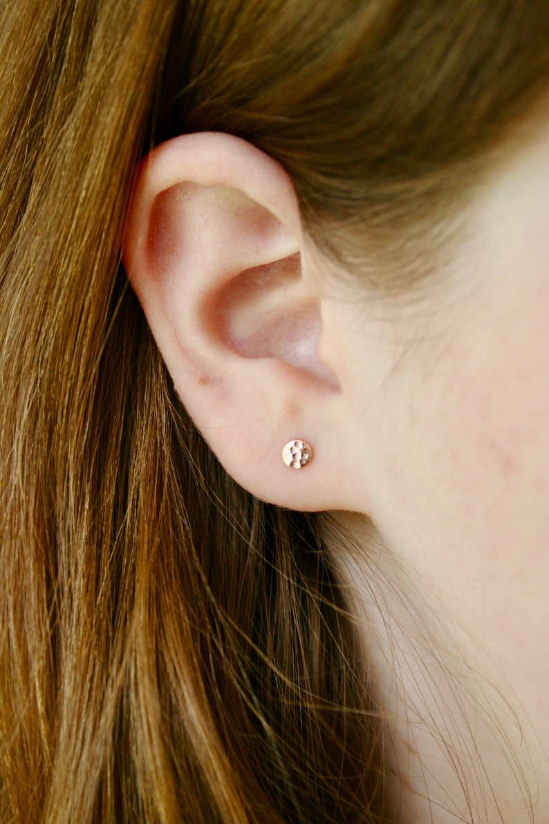 May include: A close-up of a person's ear with a small, round, hammered gold earring.