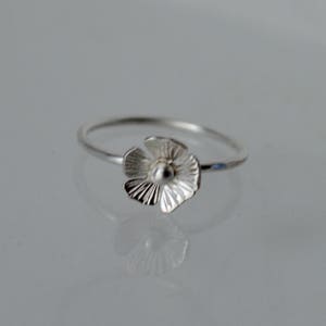 May include: A silver ring featuring a small, five-petaled flower charm. The flower has textured, radiating lines on each petal and a smooth, rounded center. The band of the ring is thin, polished, and circular, reflecting light on its metallic surface.