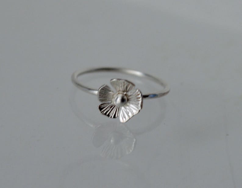 May include: A silver ring featuring a small, five-petaled flower charm. The flower has textured, radiating lines on each petal and a smooth, rounded center. The band of the ring is thin, polished, and circular, reflecting light on its metallic surface.