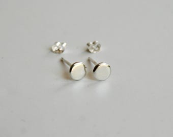 May include: A pair of silver stud earrings featuring a simple, circular, flat-faced design. The earrings include two matching silver butterfly-style earring backs positioned behind the studs, showcasing a polished metallic finish throughout.