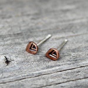 May include: A pair of copper stud earrings featuring a teardrop shape with a recessed black triangular cutout design. Each earring has a thin metal post extending from the back, designed for piercing, with a polished metallic finish on the surface.