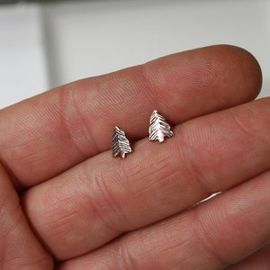 May include: A pair of small, silver-toned earrings shaped like pine trees. The earrings are on a person's hand.