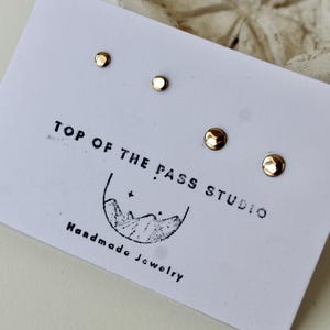 May include: Two pairs of small, round, gold-colored stud earrings attached to a white display card. The card features the black printed text TOP OF THE PASS STUDIO and Handmade Jewelry, along with a line drawing of a mountain range.