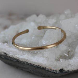 May include: A simple gold-toned bangle bracelet, made of smooth metal, rests on a bed of white crystals.
