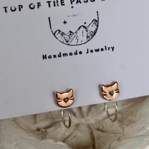 May include: Copper-coloured cat-shaped earrings with silver ear wires. The earrings are displayed on a white card with the text "TOP OF THE PASO" and "Handmade Jewelry". Each earring features a heart-shaped detail. The earrings are a cute accessory.