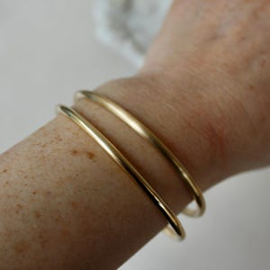 K&ouml;nnte beinhalten: Drei goldene Armreifen, die auf dem Handgelenk einer Person &uuml;bereinander liegen. Die Armreifen sind schlicht und glatt, mit einer polierten Oberfl&auml;che.