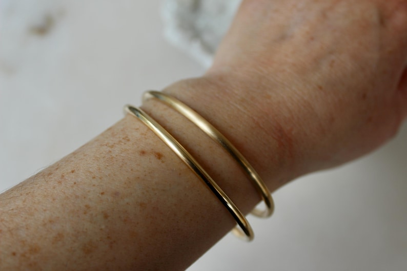 K&ouml;nnte beinhalten: Drei goldene Armreifen, die auf dem Handgelenk einer Person &uuml;bereinander liegen. Die Armreifen sind schlicht und glatt, mit einer polierten Oberfl&auml;che.