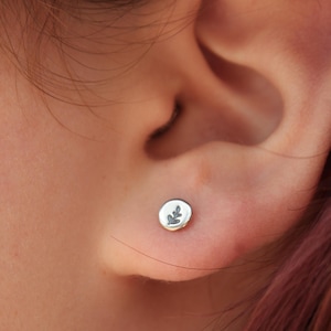May include: A small, round silver stud earring with a leaf design. The earring is set on a person's ear, with the focus on the jewelry. The earring is a simple, minimalist design.