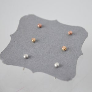 May include: A set of five small, round stud earrings in silver, gold, and rose gold. The earrings are displayed on a gray card.