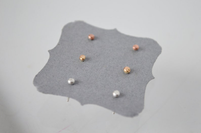 May include: A set of five small, round stud earrings in silver, gold, and rose gold. The earrings are displayed on a gray card.
