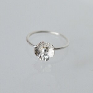 May include: A silver ring featuring a delicate flower-shaped charm with textured, radiating petals and a small, smooth spherical center. The band is thin and circular, crafted from a polished silver-toned metal with a bright, reflective finish.