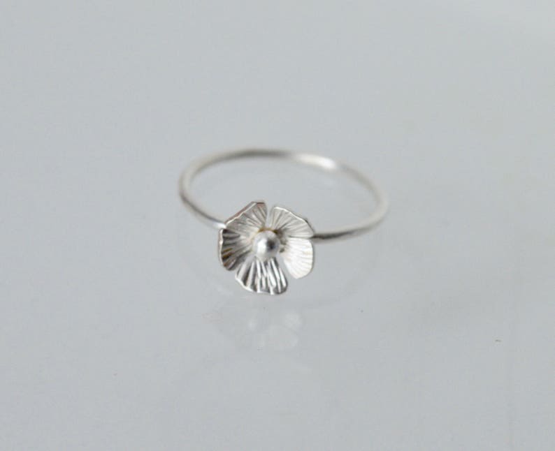 May include: A silver ring featuring a delicate flower-shaped charm with textured, radiating petals and a small, smooth spherical center. The band is thin and circular, crafted from a polished silver-toned metal with a bright, reflective finish.
