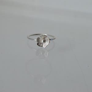 May include: A delicate silver ring featuring a small, five-petaled flower charm. The flower has a polished center and textured, radiating lines on each petal. The band is thin and circular, crafted from a smooth, reflective silver-toned metal.