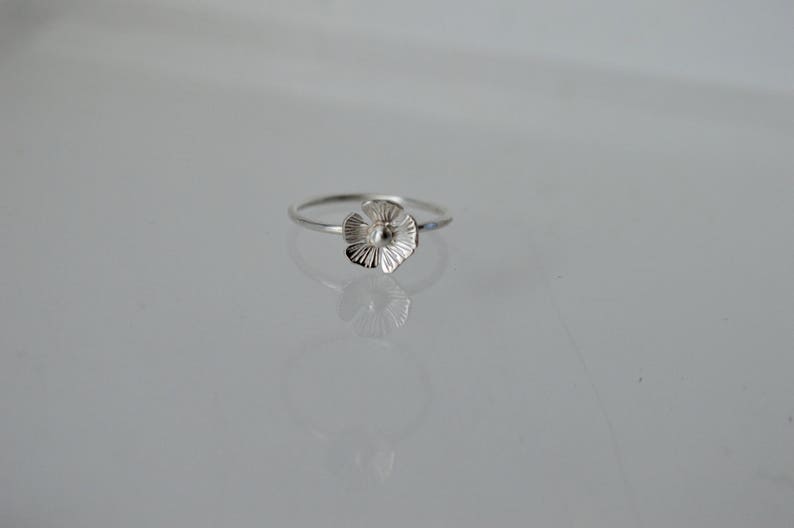 May include: A delicate silver ring featuring a small, five-petaled flower charm. The flower has a polished center and textured, radiating lines on each petal. The band is thin and circular, crafted from a smooth, reflective silver-toned metal.