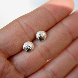 May include: Two small, round, silver-colored stud earrings. Each earring features a detailed, textured design. The earrings are displayed on a light background, showcasing their intricate patterns and metallic sheen. Jewelry.