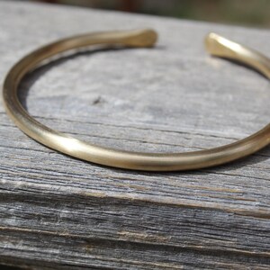 K&ouml;nnte beinhalten: Ein schlichtes goldenes Armreif-Armband aus dickem, glattem Metall. Das Armband ist offen und hat eine abgerundete Form.