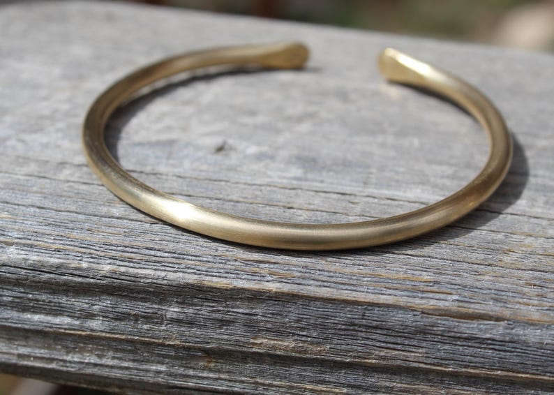 K&ouml;nnte beinhalten: Ein schlichtes goldenes Armreif-Armband aus dickem, glattem Metall. Das Armband ist offen und hat eine abgerundete Form.