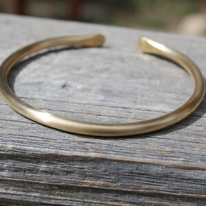 K&ouml;nnte beinhalten: Ein schlichtes goldenes Armreif-Armband aus glattem, abgerundetem Metall. Das Armband liegt auf einer Holzoberfl&auml;che.