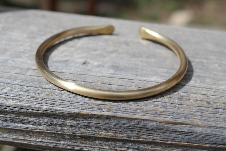 K&ouml;nnte beinhalten: Ein schlichtes goldenes Armreif-Armband aus glattem, abgerundetem Metall. Das Armband liegt auf einer Holzoberfl&auml;che.