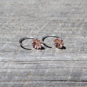 May include: A pair of silver hoop earrings with a copper-colored flower design. The earrings are displayed on a weathered wooden surface, showcasing their delicate floral detail and metallic contrast. The earrings are a jewelry item.