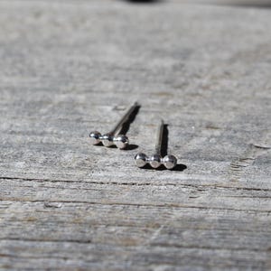 May include: A pair of silver stud earrings, each featuring three small, evenly spaced spheres. The earrings are displayed on a weathered, gray wooden surface, with natural light casting shadows. The minimalist design offers a simple, elegant aesthetic.