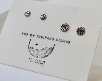 May include: Four silver stud earrings with a sunburst design are displayed on a white card. The card has a stamp that reads "TOP OF THE PASS STUDIO" and "MADE IN COLORADO". The earrings vary in size, with two pairs of matching designs.