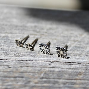 May include: Four silver stud earrings with a geometric, sunburst design. The earrings are square-shaped with a textured surface and are displayed on a weathered wooden surface. The earrings are small and delicate.