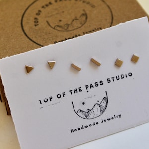May include: A set of gold-colored stud earrings in various geometric shapes, including triangles, rectangles, and squares, displayed on a white card. The card is printed with "TOP OF THE PASS STUDIO" and a mountain logo. The earrings are presented with a brown cardboard box.