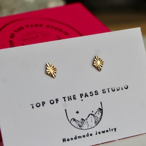 May include: A pair of small, gold-colored stud earrings with a sunburst design. The earrings are displayed on a white card with the text "TOP OF THE PASS STUDIO" and a mountain logo. A red box is in the background.