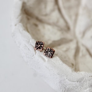 May include: Two square copper earrings with a carved starburst design. The earrings are displayed on a textured, off-white surface, creating a contrast between the warm metal and the neutral background. These earrings are a stylish accessory.
