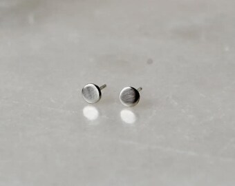 May include: A pair of small, round, silver stud earrings. Each earring has a brushed metal finish and a post for pierced ears. The earrings are displayed on a light-colored, marble-like surface.