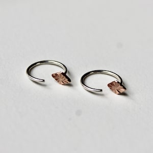 May include: A pair of silver-colored hoop earrings, each with a small, rose-gold colored diamond-shaped accent. The earrings are open at one end and rest on a white surface. The rose-gold accents have a textured, patterned surface.