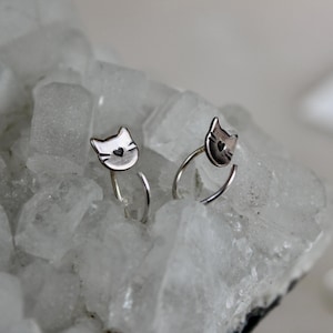May include: A pair of silver cat-shaped earrings. Each earring features a cat face with a heart detail and curved post. The earrings are set against a white, crystalline background. The cat face has a heart detail.