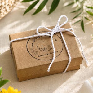 May include: A rectangular brown cardboard gift box tied with a white twisted cord bow. The lid features a circular black ink stamp with the text Top of the Page Studio and Handmade Jewelry, accompanied by a line drawing of mountains and stars.