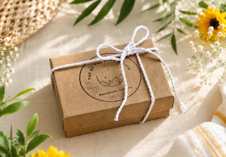 May include: A rectangular brown cardboard gift box tied with a white twisted cord bow. The lid features a circular black ink stamp with the text Top of the Page Studio and Handmade Jewelry, accompanied by a line drawing of mountains and stars.