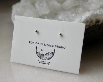 May include: Two small, silver stud earrings presented on a white card. The card is printed with "TOP OF THE.PASS STUDIO" and a mountain logo, with "HANDMADE IN COLORADO" below. The earrings are a minimalist design.
