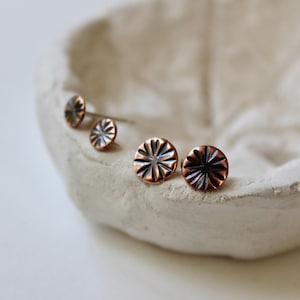 May include: Two pairs of round copper stud earrings featuring a sunburst or flower-like pattern with dark recessed lines radiating from the center. The metal has a warm, reddish-brown tone with a polished finish on the raised surfaces.