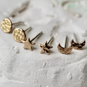 May include: A set of six gold-toned stud earrings featuring a hammered texture. The collection includes two circular studs, two five-pointed star studs, and two crescent moon studs, each equipped with a thin metal post for secure attachment.