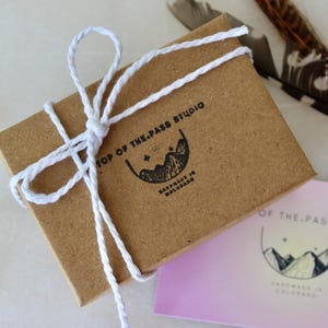 K&ouml;nnte beinhalten: Eine kleine braune Schachtel, die mit wei&szlig;em Garn gebunden ist. Die Schachtel hat ein Etikett mit der Aufschrift "Top of the Pass Studio Handmade in Colorado".