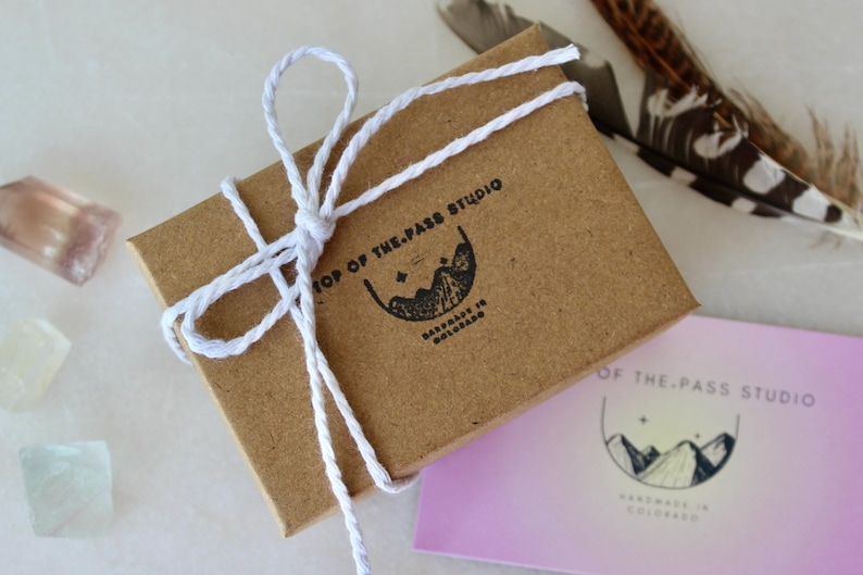 K&ouml;nnte beinhalten: Eine kleine braune Schachtel, die mit wei&szlig;em Garn gebunden ist. Die Schachtel hat ein Etikett mit der Aufschrift "Top of the Pass Studio Handmade in Colorado".