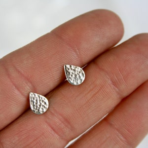 May include: A pair of small, silver teardrop-shaped stud earrings. The earrings have a hammered texture and are displayed on a person's hand. The earrings are a simple, elegant design.