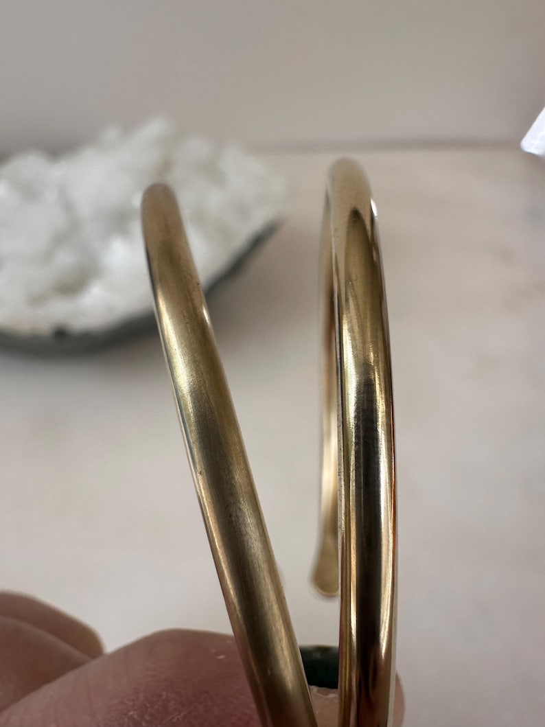 K&ouml;nnte beinhalten: Zwei goldfarbene Metallarmb&auml;nder, eines etwas breiter als das andere, mit einer glatten, polierten Oberfl&auml;che.