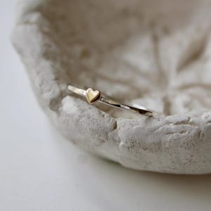 May include: A delicate ring with a small gold heart set on a silver band. The ring is displayed on a textured, off-white surface, showcasing its simple elegance. This piece is a charming addition to any jewelry collection.