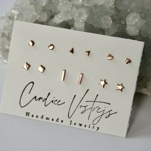 May include: A set of nine gold-toned stud earrings on a white card. The earrings are in various shapes, including circles, squares, triangles, stars, and hearts. The card has the text "Candice Ostryjs Handmade Jewelry".