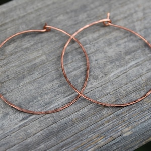 May include: Two copper-colored hoop earrings with a hammered texture. The earrings are circular and have a simple, minimalist design. They are resting on a weathered, gray wooden surface.