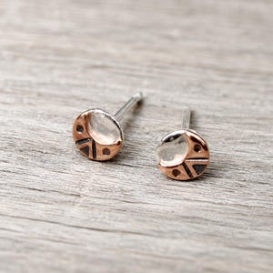 May include: A pair of small stud earrings featuring a circular design with a copper-toned finish. Each earring has a smooth, rounded top section and a lower portion with dark, recessed geometric lines and small circular indentations.